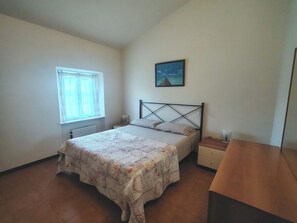 A bedroom with a bed, a nightstand, a window with curtains, a picture on the wall, and a wooden desk.