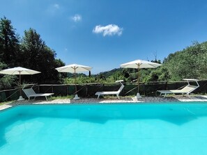 Una piscina con sillas de descanso y sombrillas, rodeada de un muro de piedra y vegetación.