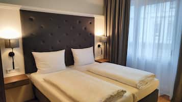 Cama doble con cabecera capitonada, dos mesitas de noche con lámparas y vistas a una ventana con cortinas.