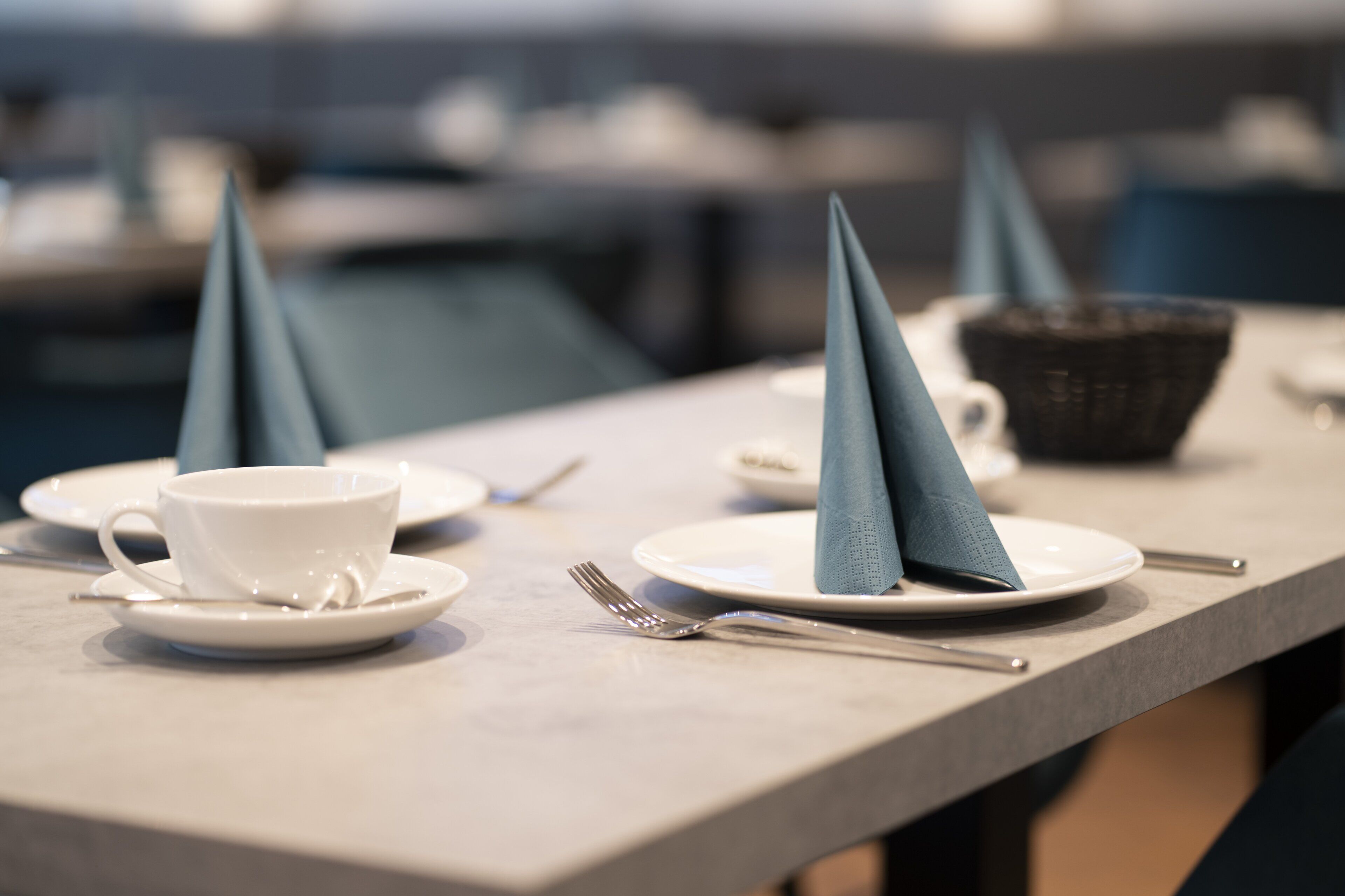 Una mesa de comedor con una taza blanca, un plato bajo y un tenedor. Hay servilletas dobladas en forma de conos sobre los platos.