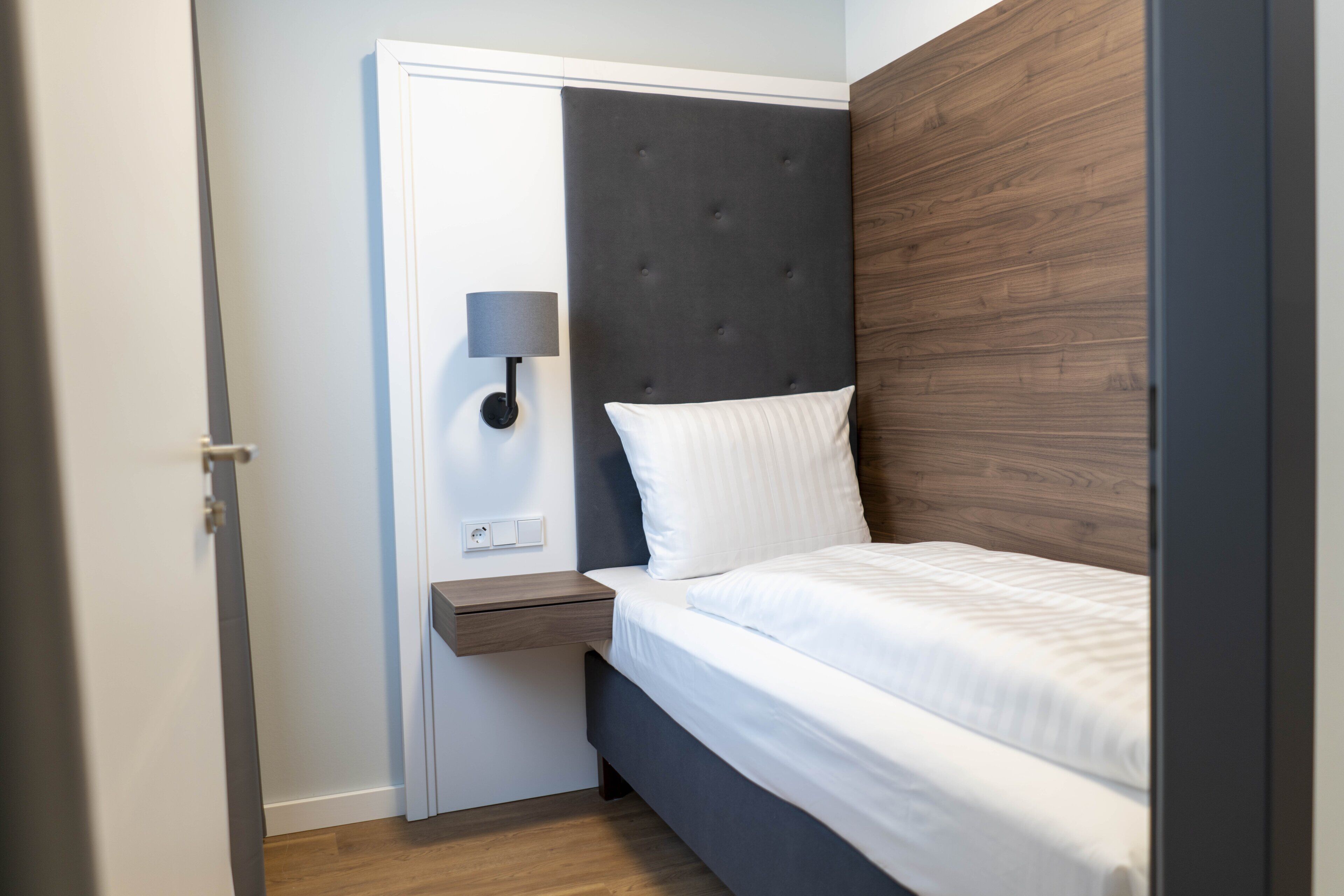 A bedroom with a bed, a nightstand, a wall-mounted lamp, and a wooden headboard.
