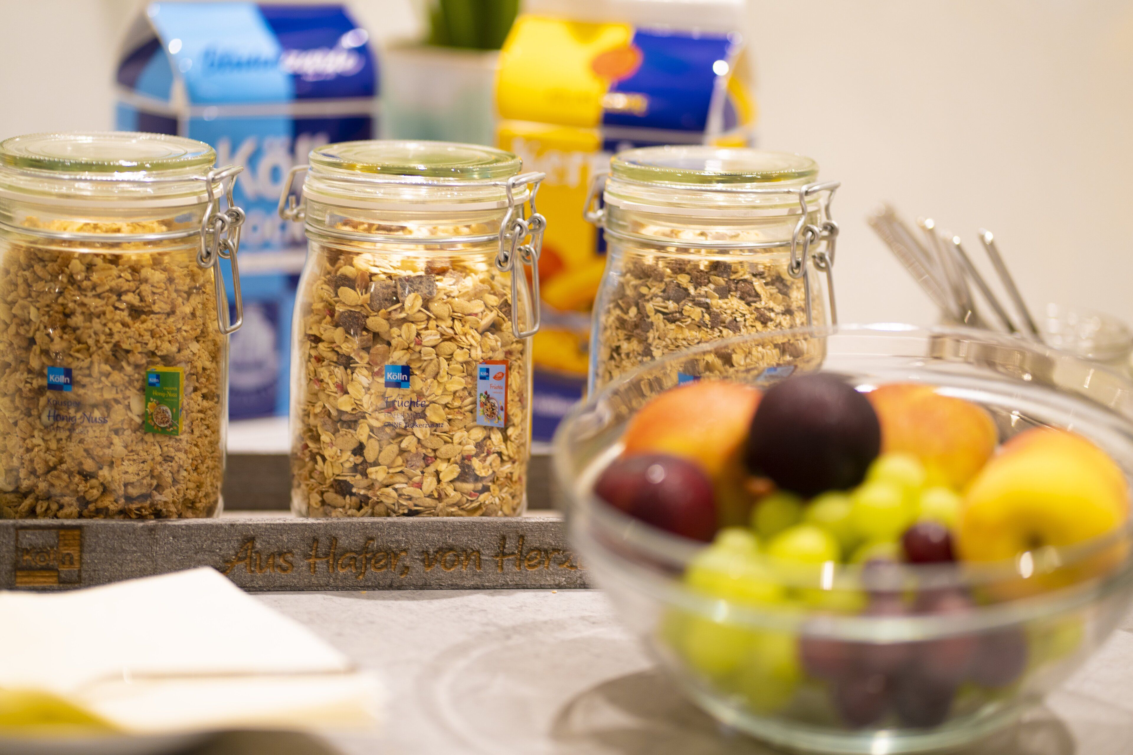 Drei Glaskolben mit Müsli und Metallverschlüssen, eine Schüssel mit gemischten Früchten und ein blau-gelb verpacktes Produkt im Hintergrund.