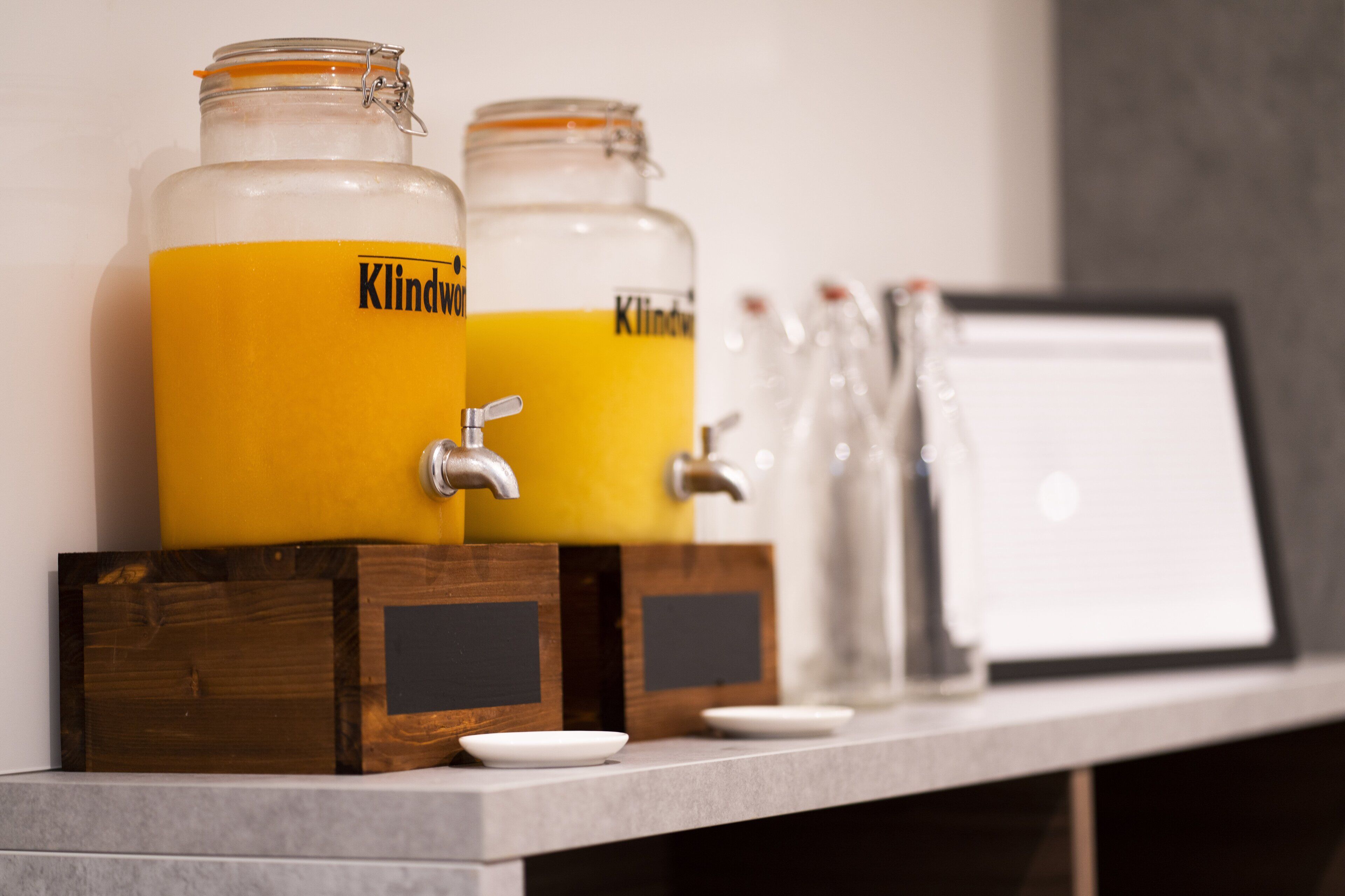 Due erogatori in vetro capienti con liquido giallo, etichettati 'Kindway', su una base di legno con un rubinetto.
