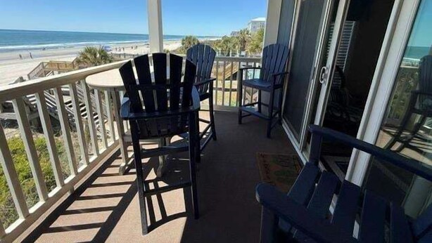 A balcony with chairs and a table, overlooking a beach and ocean.