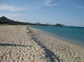 Une vaste plage de sable fin, bordée d’une eau d’un bleu limpide et dominée par des montagnes lointaines.