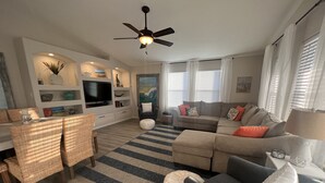 A modern living room with a sofa, ottoman, and a dining area with wicker chairs. - Winds of Change - Easy Beach Access (Bolivar Peninsula)