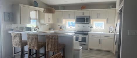Een keuken met witte kastjes, een centraal eiland met barkrukken, een magnetron, fornuis en een spoelbak.