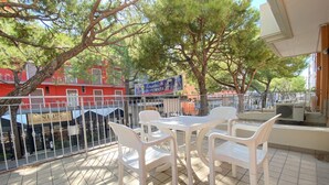 A terrace with a table and chairs, a view of a river, and a building with a sign in the background. - Appartamento Adriana 2 - Carraro Immobiliare Jesolo (Lido di Jesolo)