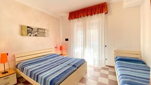 A bedroom with two beds, a nightstand, a lamp, a radiator, and a window with curtains. - Appartamento Adriana 2 - Carraro Immobiliare Jesolo (Lido di Jesolo)