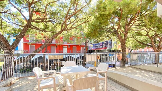 An outdoor seating area with white plastic chairs and tables, surrounded by a metal fence, with trees and a red building in the background. - Appartamento Adriana 2 - Carraro Immobiliare Jesolo (Lido di Jesolo)