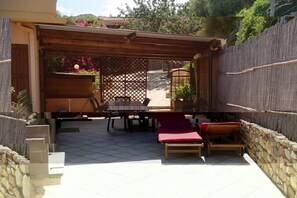 Um pátio externo com pérgola de madeira, uma mesa, duas poltronas de descanso e uma parede de pedra.