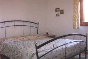 Uma cama de solteiro com estrutura de metal, uma mesa de cabeceira com abajur e uma janela com cortinas.