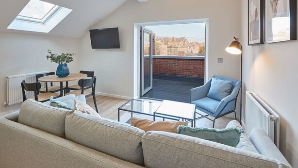 A modern living room with a sofa, a coffee table, a blue chair, a dining table with chairs, a TV mounted on the wall, and a large window with a view of buildings. - Host & Stay | Skyline Apartment (Whitby)