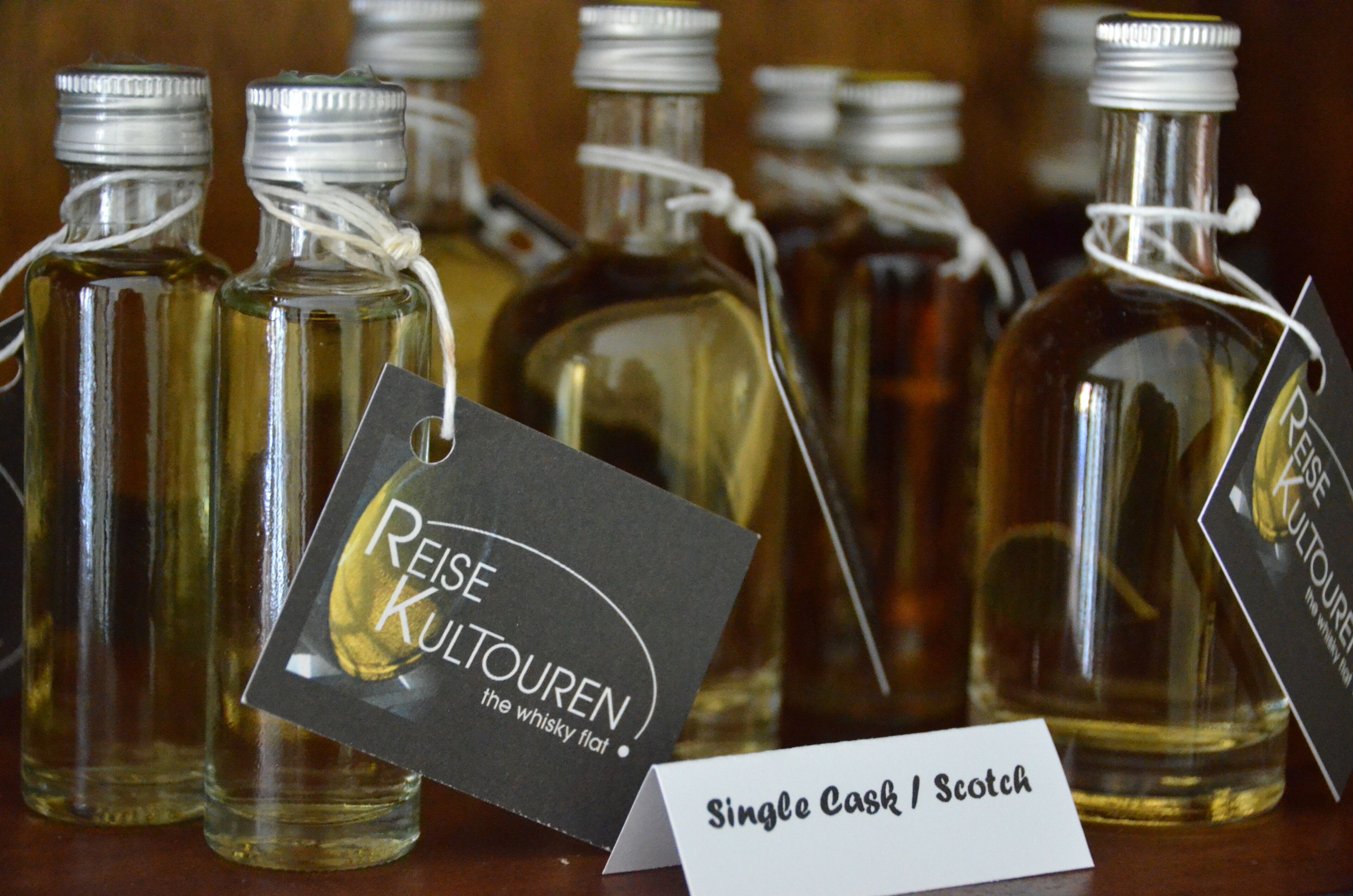 Several glass bottles with metal caps, each with a tag. One tag reads 'Reise Kulturen the whisky flat.' Another tag says 'Single Cask / Scotch.'