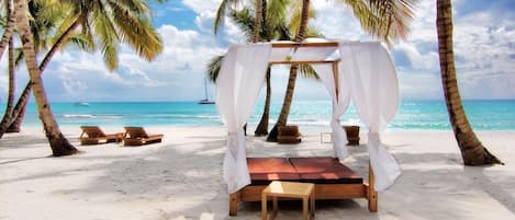 Un paisaje playero con una cama con dosel, sillas de madera para relajarse y palmeras.