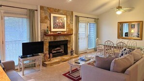 Ein Wohnzimmer mit Kamin, Fernseher, einer Couch, einem Esstisch mit Stühlen und einem Deckenventilator.