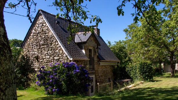 Una casa de piedra con techo a dos aguas, rodeada de abundante vegetación y plantas con flores.