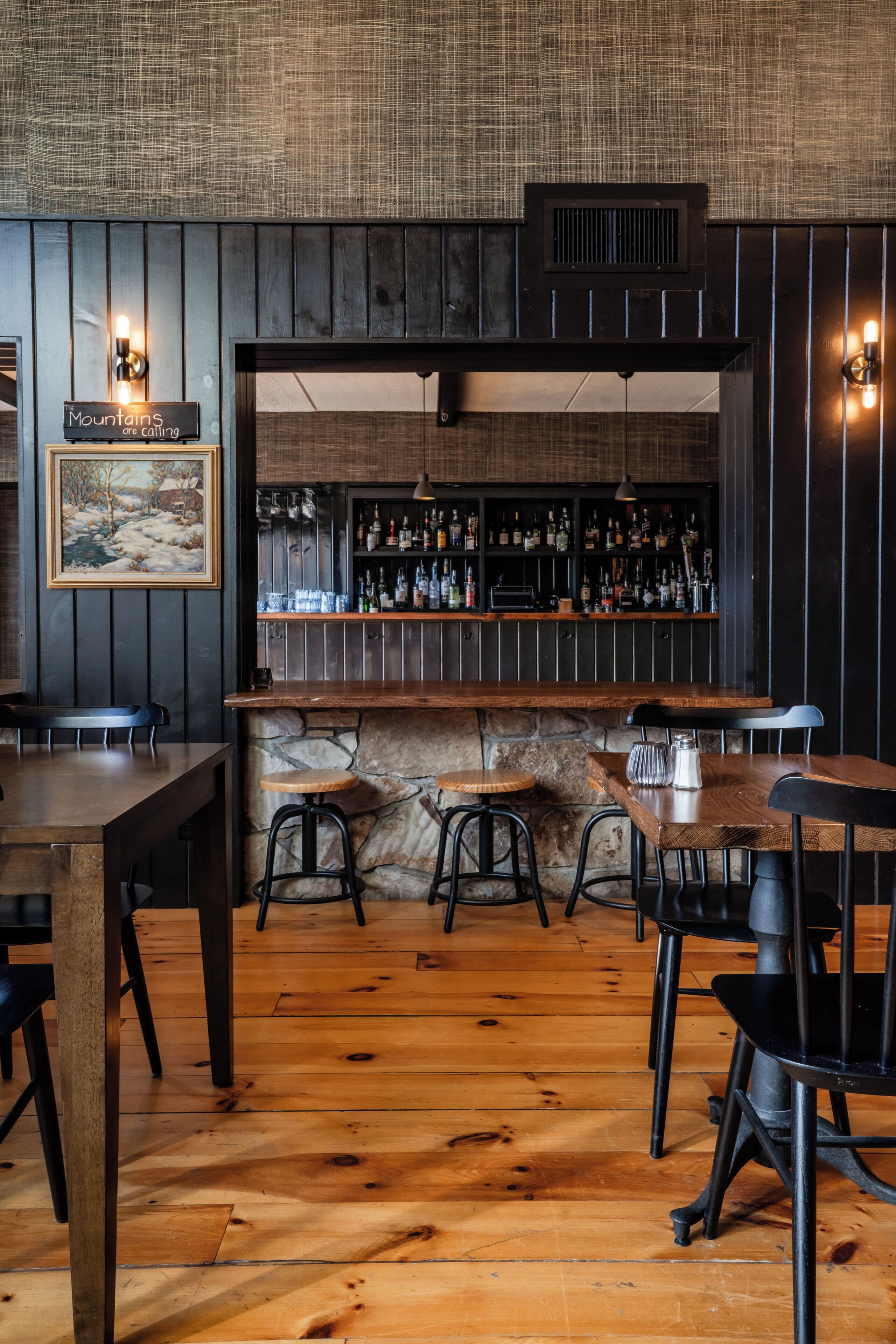 A bar area with wooden flooring, bar stools, a stone counter, and a wall-mounted shelf with bottles.