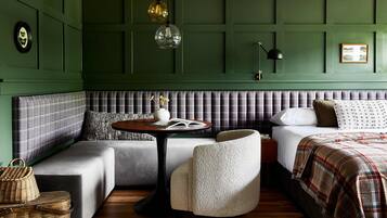 A room with a green paneled wall, a grey sofa, a white armchair, a bed with a plaid blanket, and a small round table with a book and vase.