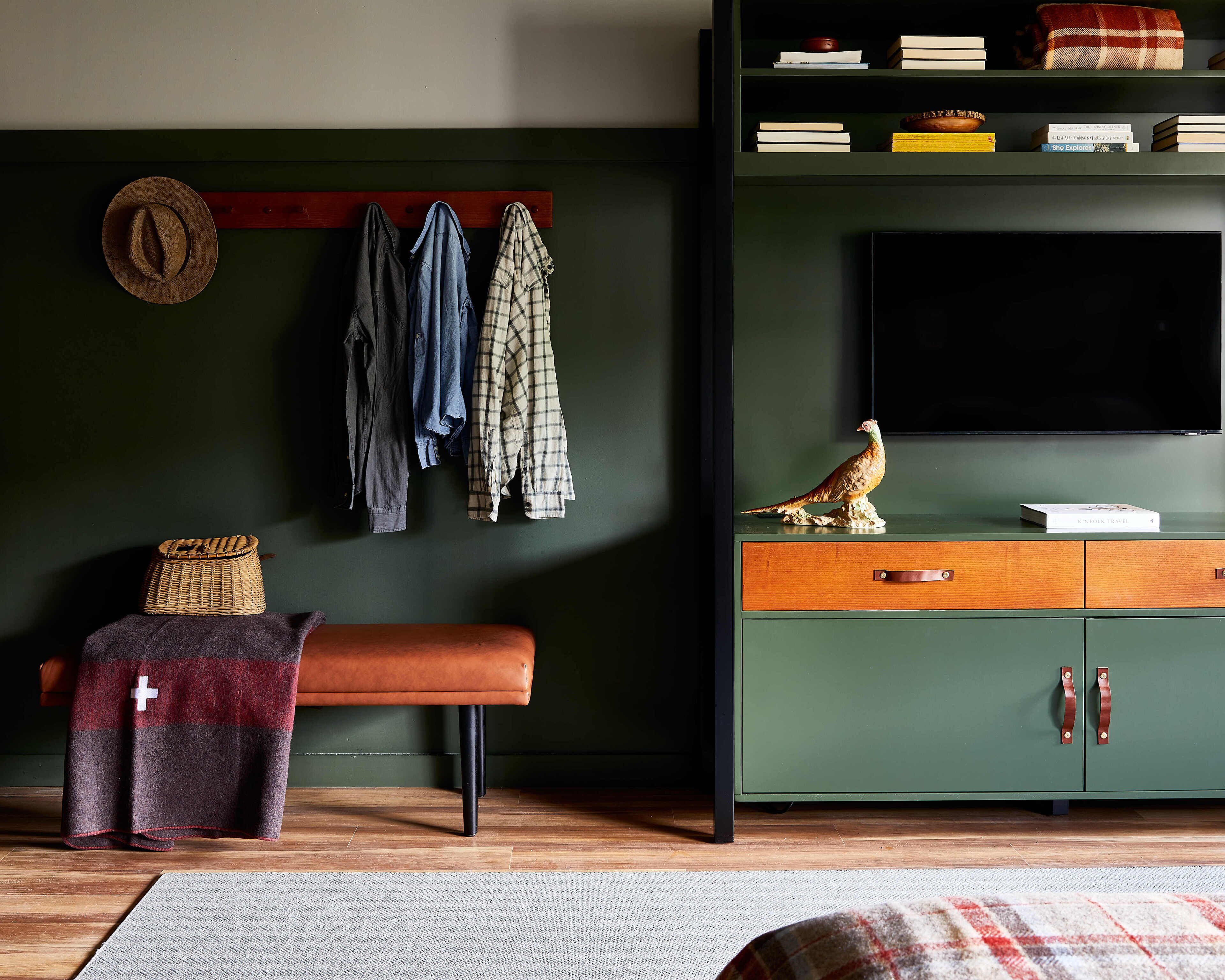 Quarto com parede verde, um banco de madeira, um chapéu e um cabideiro com jaquetas.