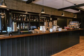 A bar area with a counter, shelves of bottles, a coffee station, and hanging lights.