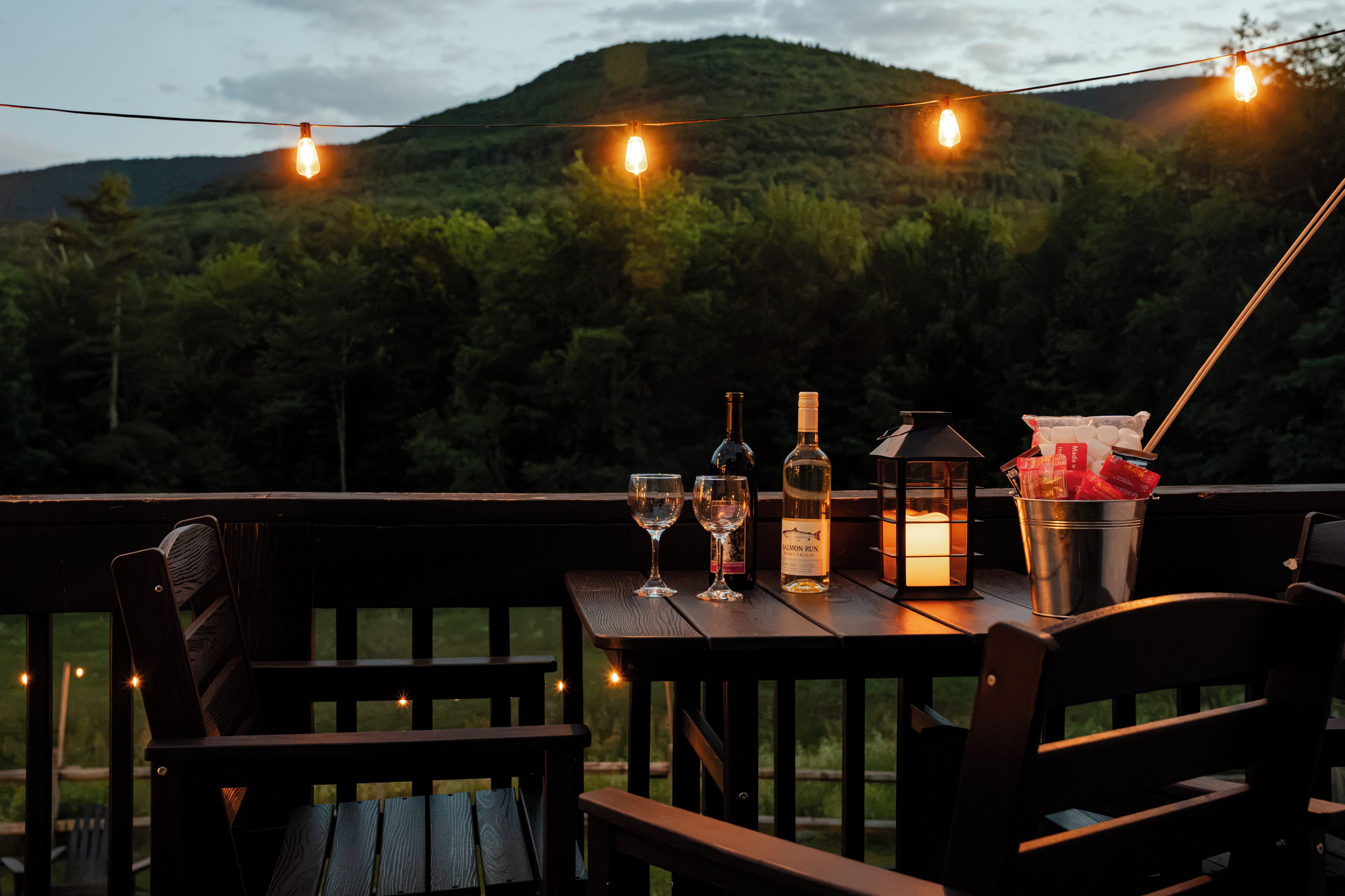 A wooden deck with a table set for dining, featuring a bottle of wine, glasses, and a lantern.
