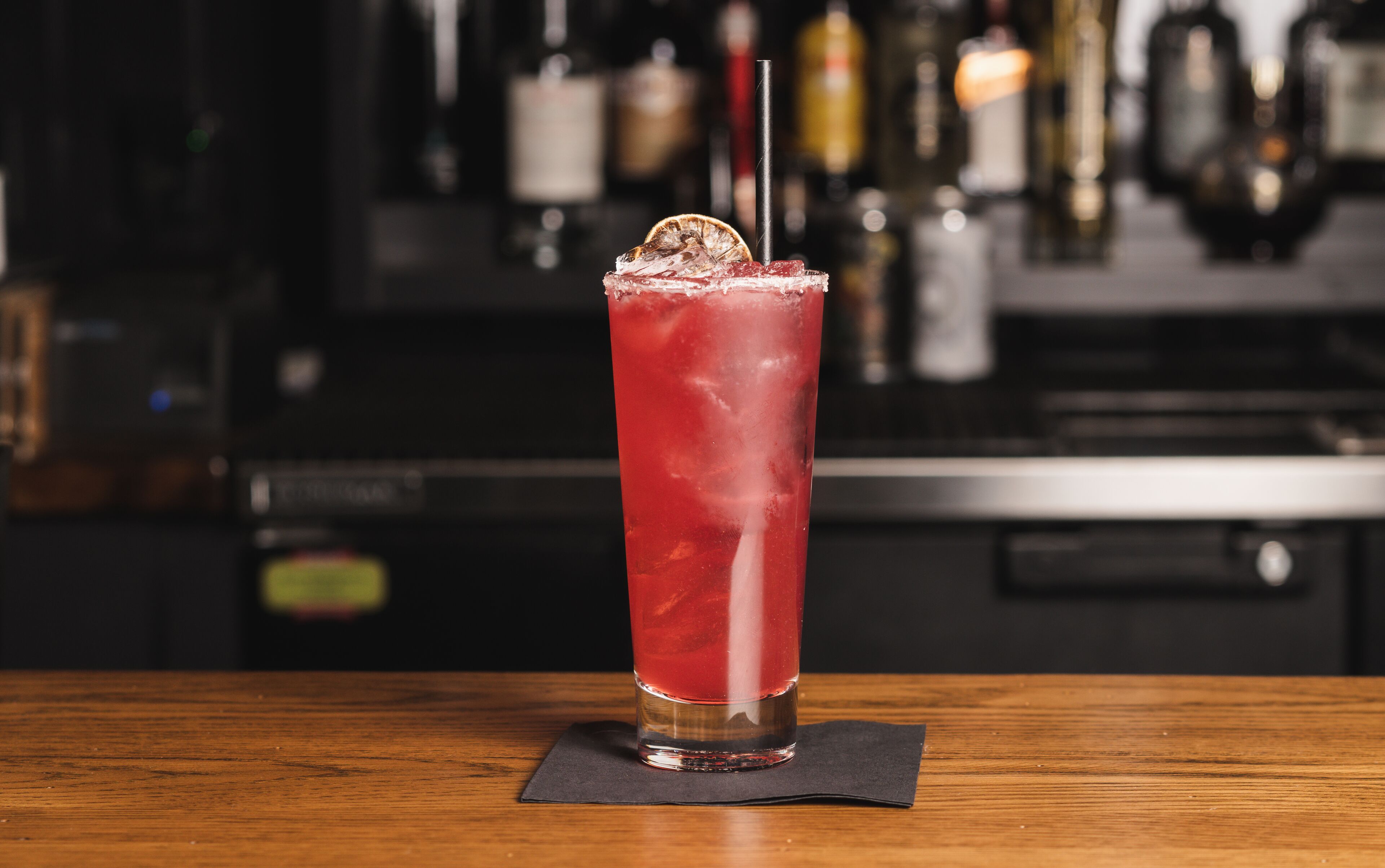 A tall glass of red beverage with a straw and a slice of orange on a wooden bar counter.