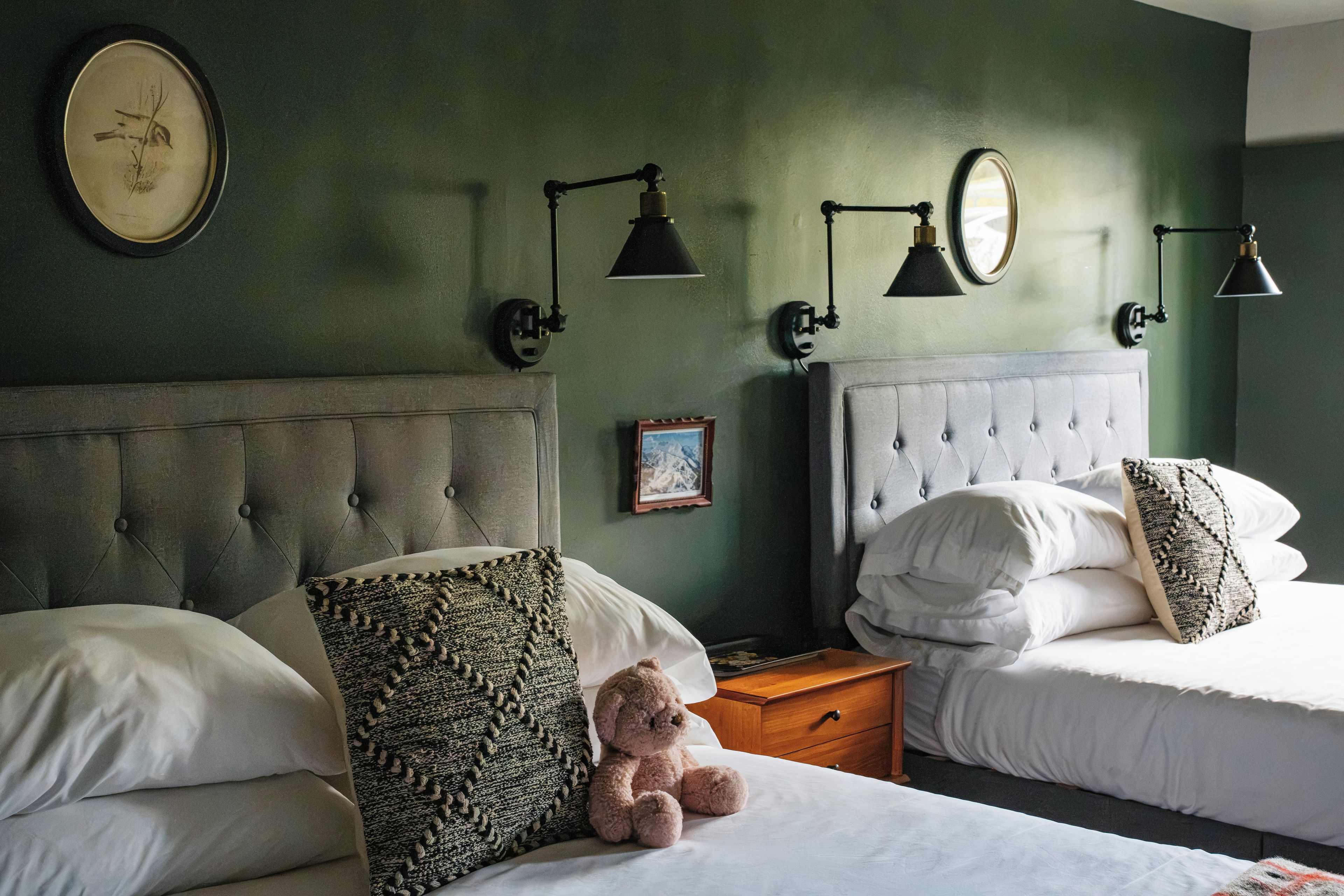 A hotel room with two beds, a nightstand, a teddy bear, and wall-mounted lamps.
