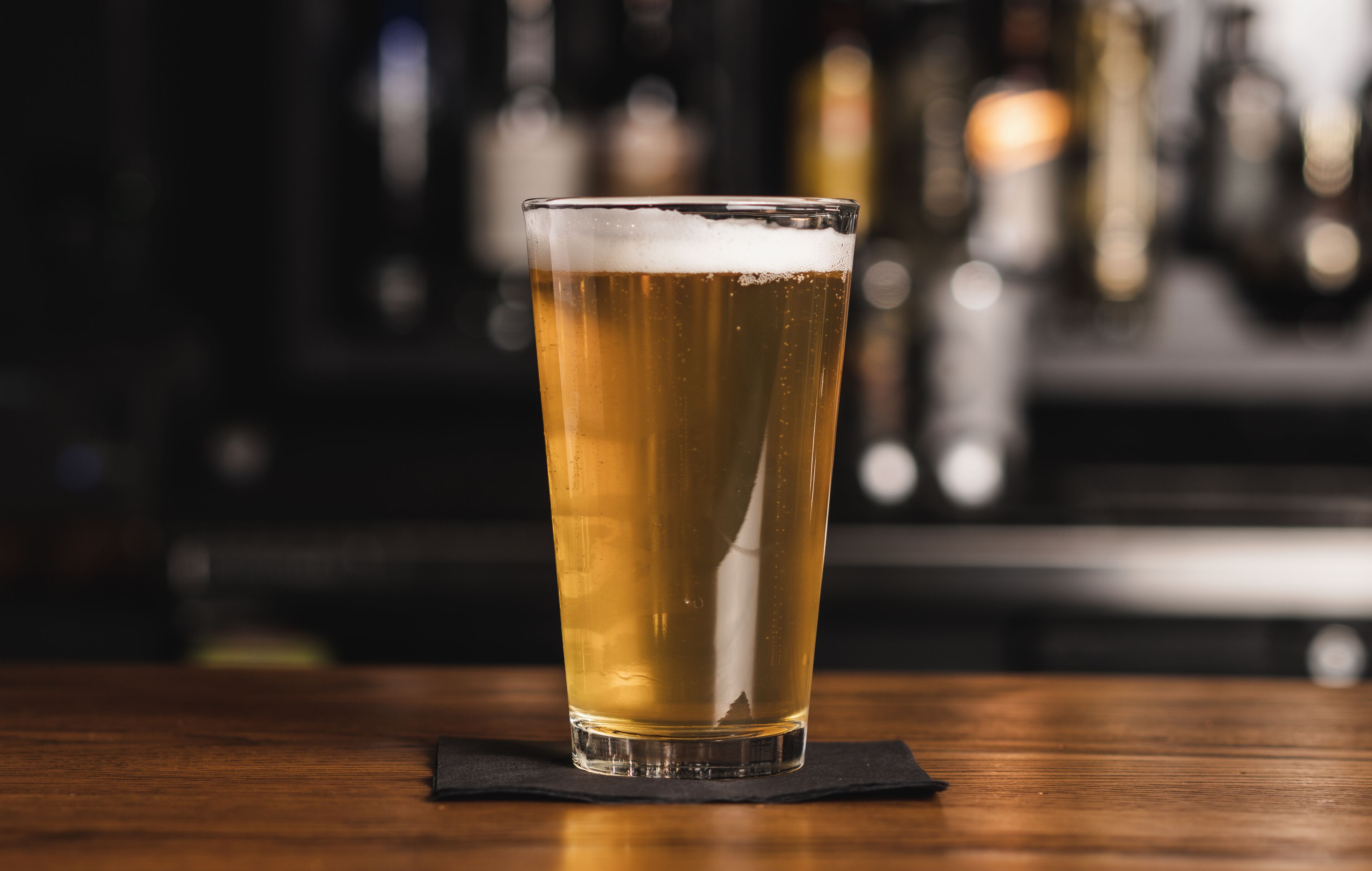 A tall glass of beer on a wooden table with a blurred background of bottles.