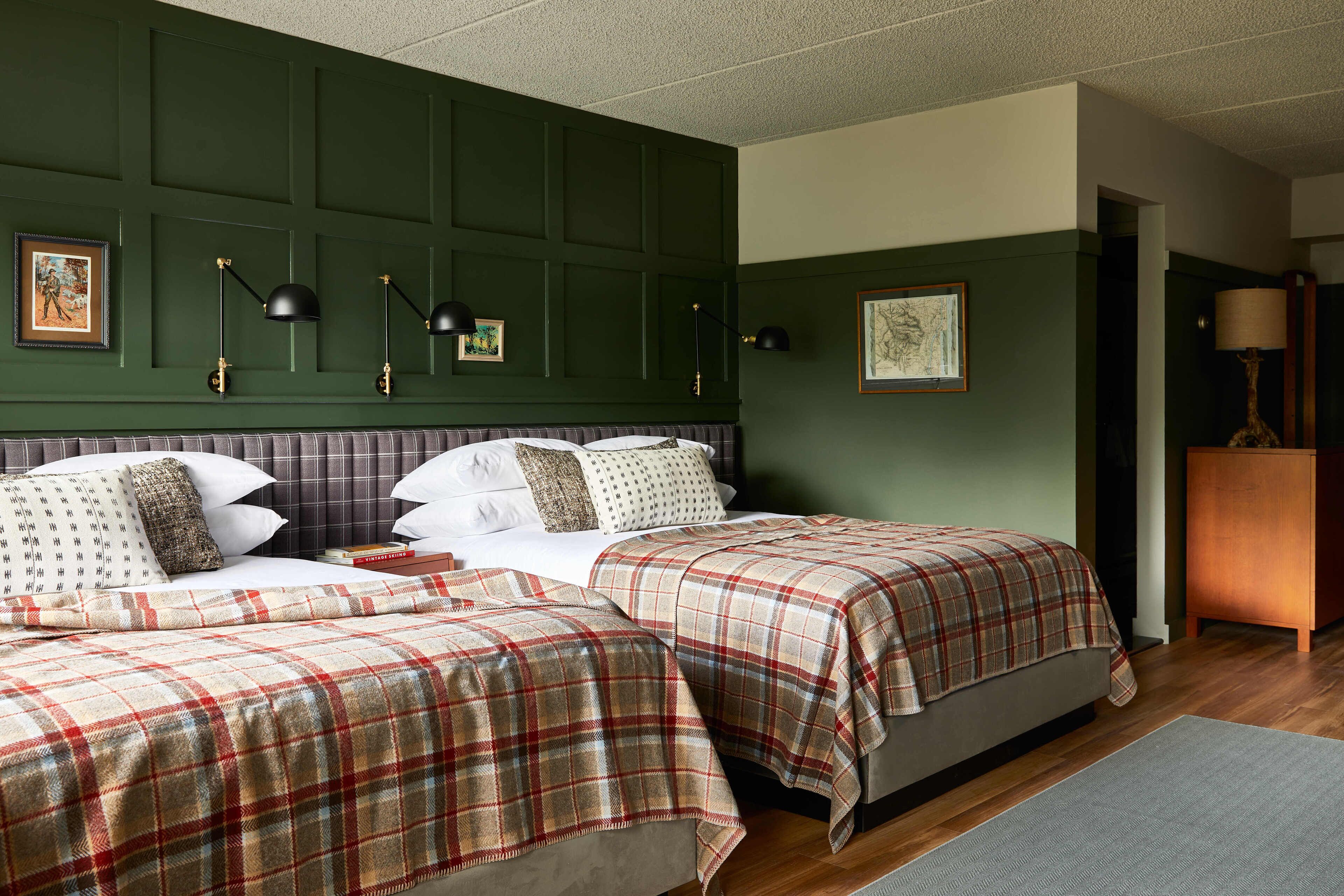 A room with two beds, each with a plaid bedspread, a headboard, and a bedside table with a lamp.