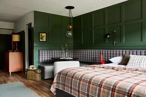 A bedroom with a plaid bedspread, a green paneled headboard, a bedside table with a lamp, and a wicker basket.