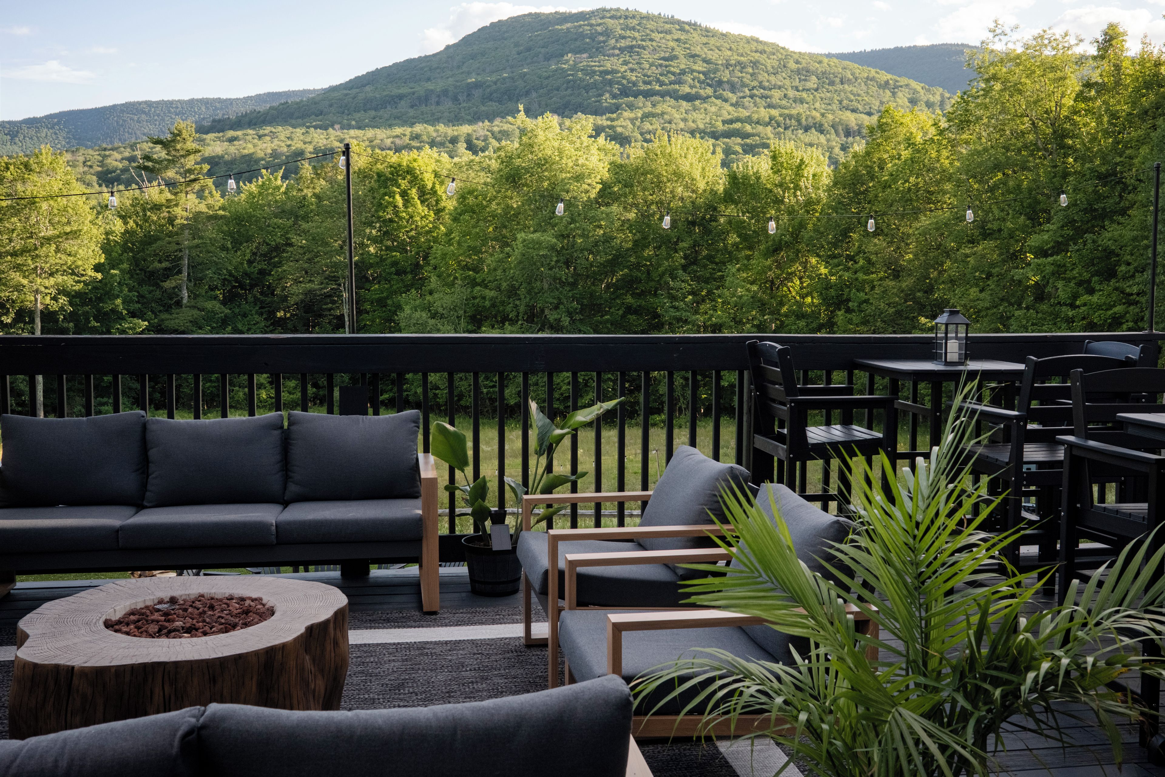 A deck with outdoor furniture, including a sofa and chairs, surrounded by greenery and a view of a forested hill.