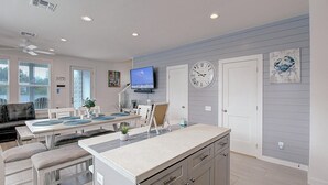 Une cuisine moderne avec un îlot central, un coin repas avec une table et des chaises blanches, un téléviseur à écran plat fixé au-dessus de l’îlot et une horloge au mur.