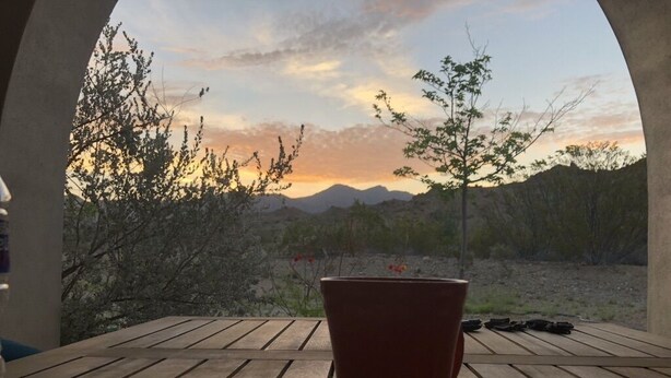 Ein Holztisch mit einem roten Topf und einem Stein, mit Blick auf eine Wüstenlandschaft mit vereinzelter Vegetation und Bergen in der Ferne.