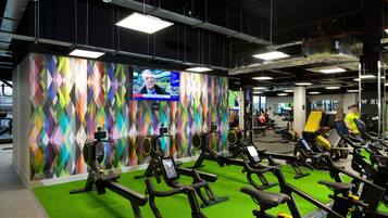 Ein Fitnessraum mit Ergometer, grünem Boden und einer farbenfrohen Wand mit Bildschirm.