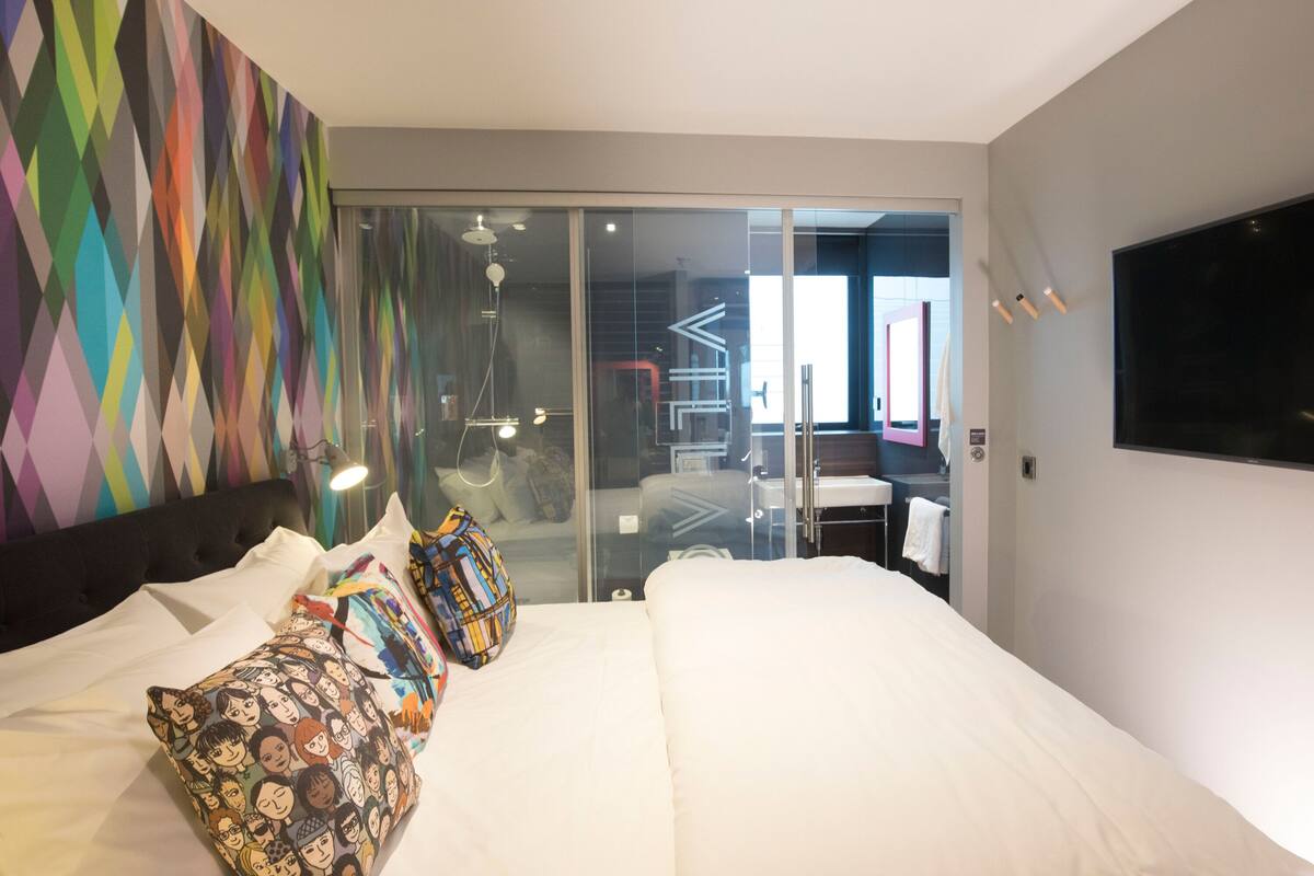 Une chambre d’hôtel moderne avec un grand lit, des oreillers colorés, un téléviseur à écran plat et une porte-fenêtre coulissante donnant sur la salle de bain.