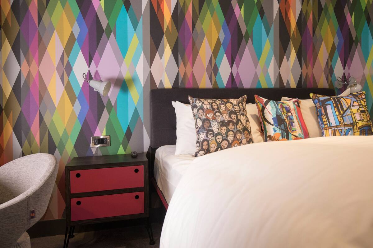 Une chambre d’hôtel avec un mur aux motifs géométriques colorés, un lit avec de nombreux oreillers, une table de chevet avec une lampe et une chaise.