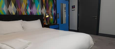 A hotel room with a bed, a desk, a chair, a lamp, a wardrobe, and a door.