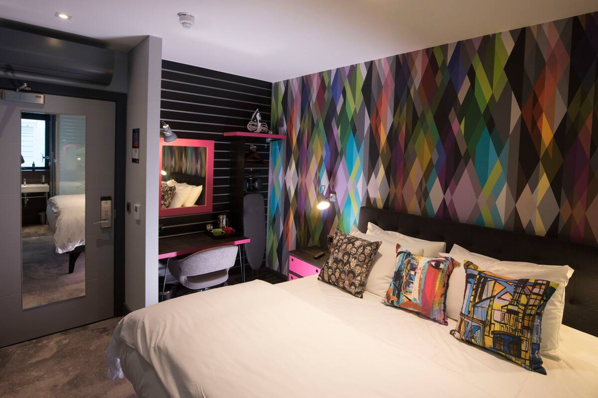 Une chambre d’hôtel moderne, équipée d’un lit, d’un bureau, d’une chaise, d’un miroir et ornée d’un motif géométrique coloré sur le mur.