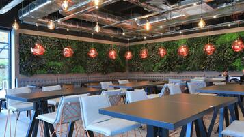 A modern dining area with tables and chairs, a green wall with hanging red lights, and a ceiling with exposed pipes and lights.