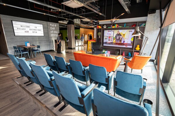 A modern lounge area with blue chairs, an orange seating area, and a large screen displaying a program. - Village Hotel Southampton Eastleigh (Eastleigh)