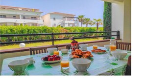 A breakfast table with fruits, pastries, and drinks, overlooking a landscaped garden and buildings.