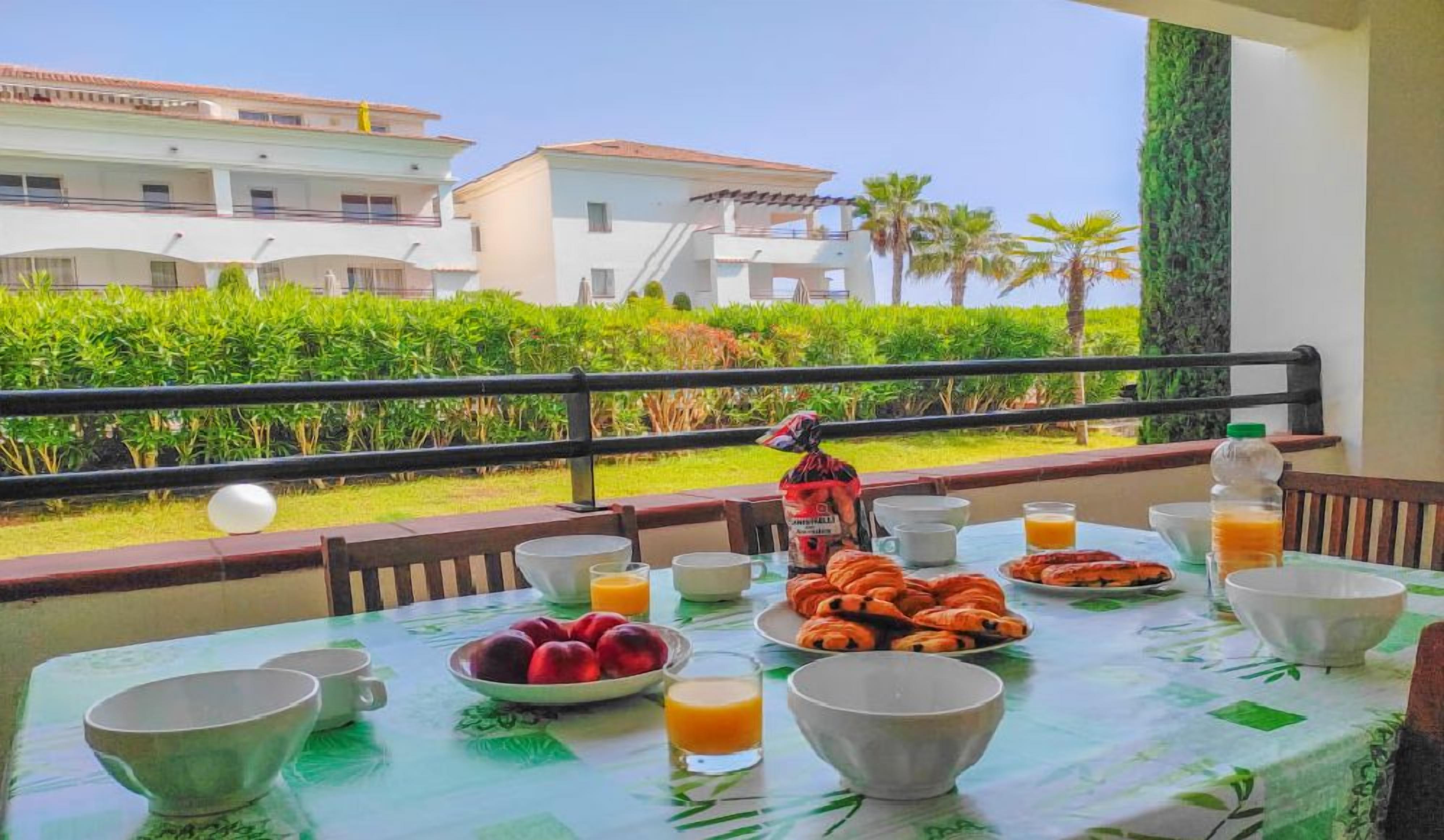 Taula de petit esmorzar amb fruita, pastissos i begudes, amb vista a un jardí ben cuidat i edificis.