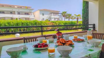 Una mesa de desayuno con frutas, pasteles y bebidas, con vistas a un jardín ajardinado y edificios.
