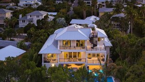 Ett stort hus med flera våningar och en swimmingpool, omgivet av frodig grönska och med utsikt över havet vid solnedgången.