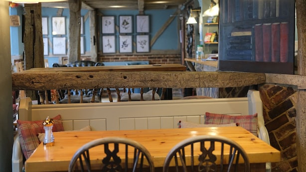 A cozy restaurant interior with exposed wooden beams, a wooden table with chairs, and a bench with plaid cushions.