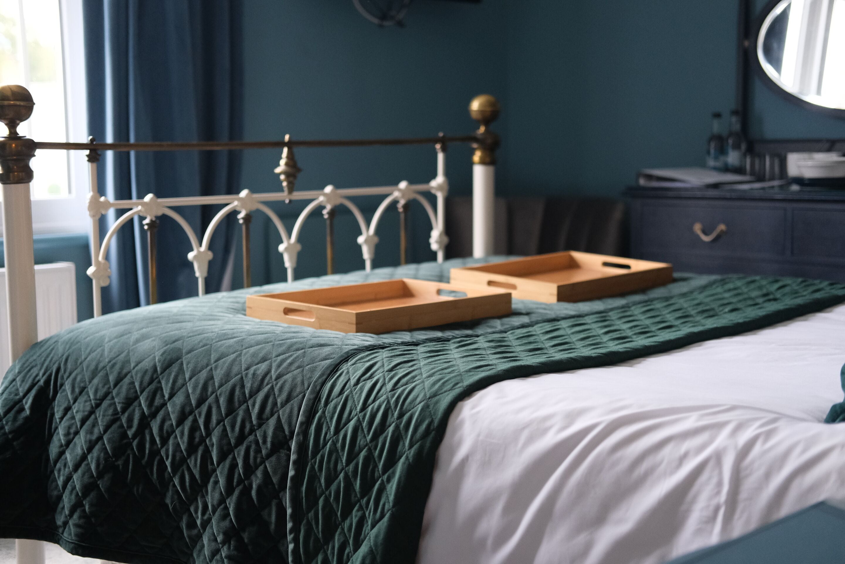 Una cama bien hecha con una manta verde acolchada y una colcha blanca. Hay una mesita de madera sobre la cama, y un aparador con un espejo y algunos objetos encima.