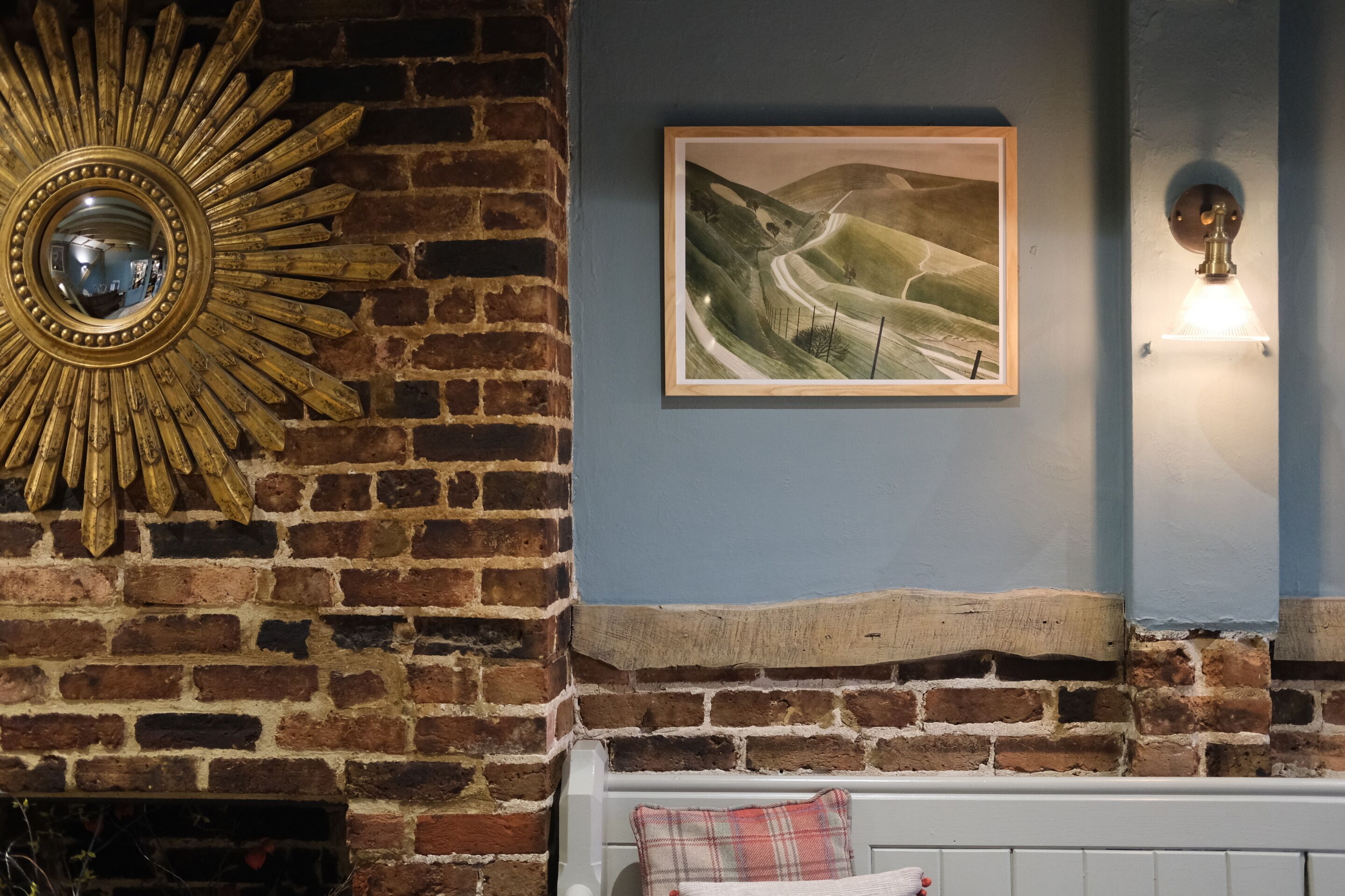 A room with a brick fireplace, a sunburst mirror, a framed landscape painting, and a white bench with a plaid pillow.