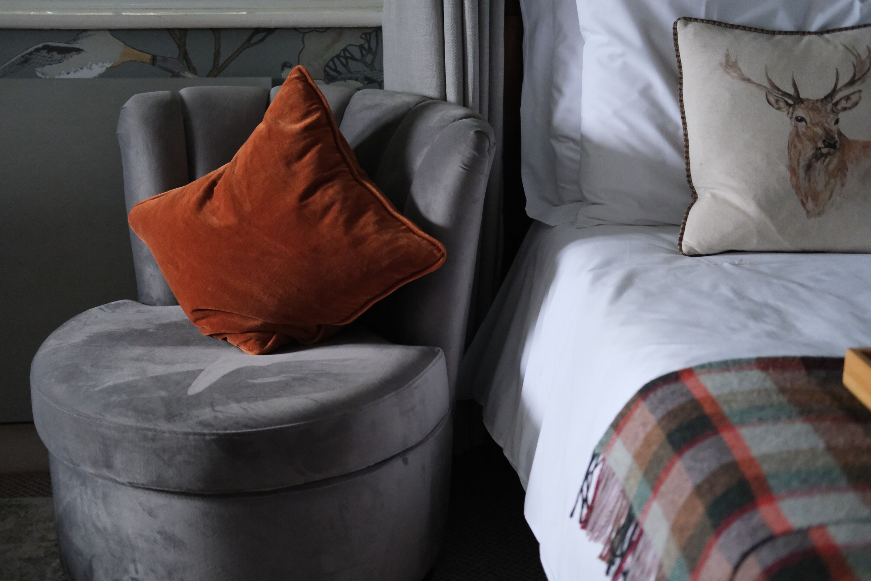 A grey ottoman with an orange cushion, a white pillow with a deer design, and a plaid blanket on a bed.