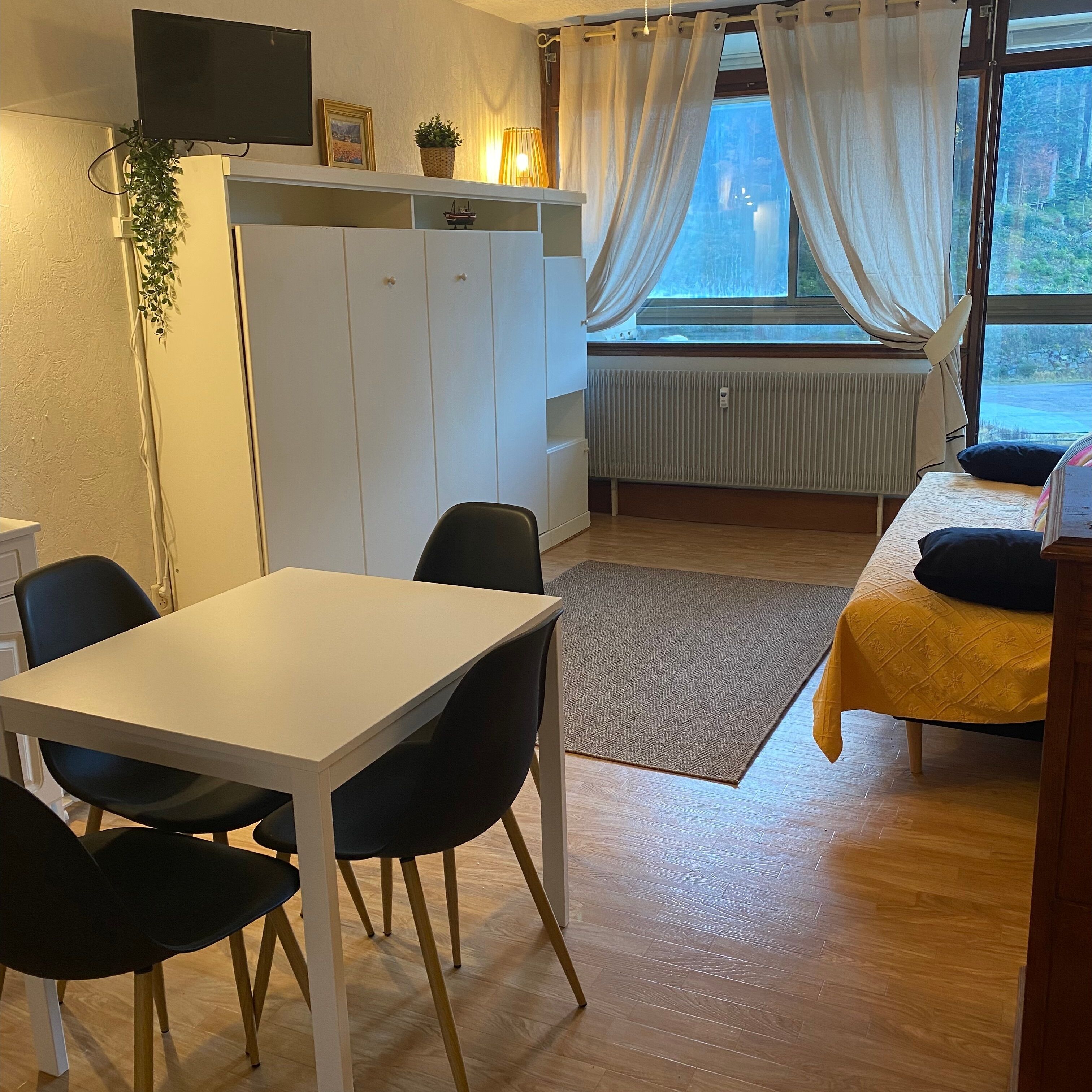 A small, well-lit room with a white table and chairs, a bed with a yellow bedspread, a radiator, and a window with curtains.